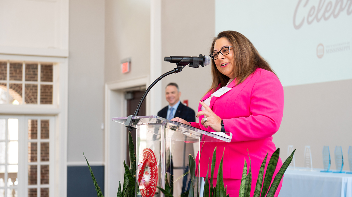 Kathleen Grigsby in a bright pink blazer speaks at a podium bearing the University of Mississippi seal during an indoor event. A projection screen displaying the word "Celebr" is partially visible in the background, along with a man in a suit seated behind her and a "School of Education" banner to the right.